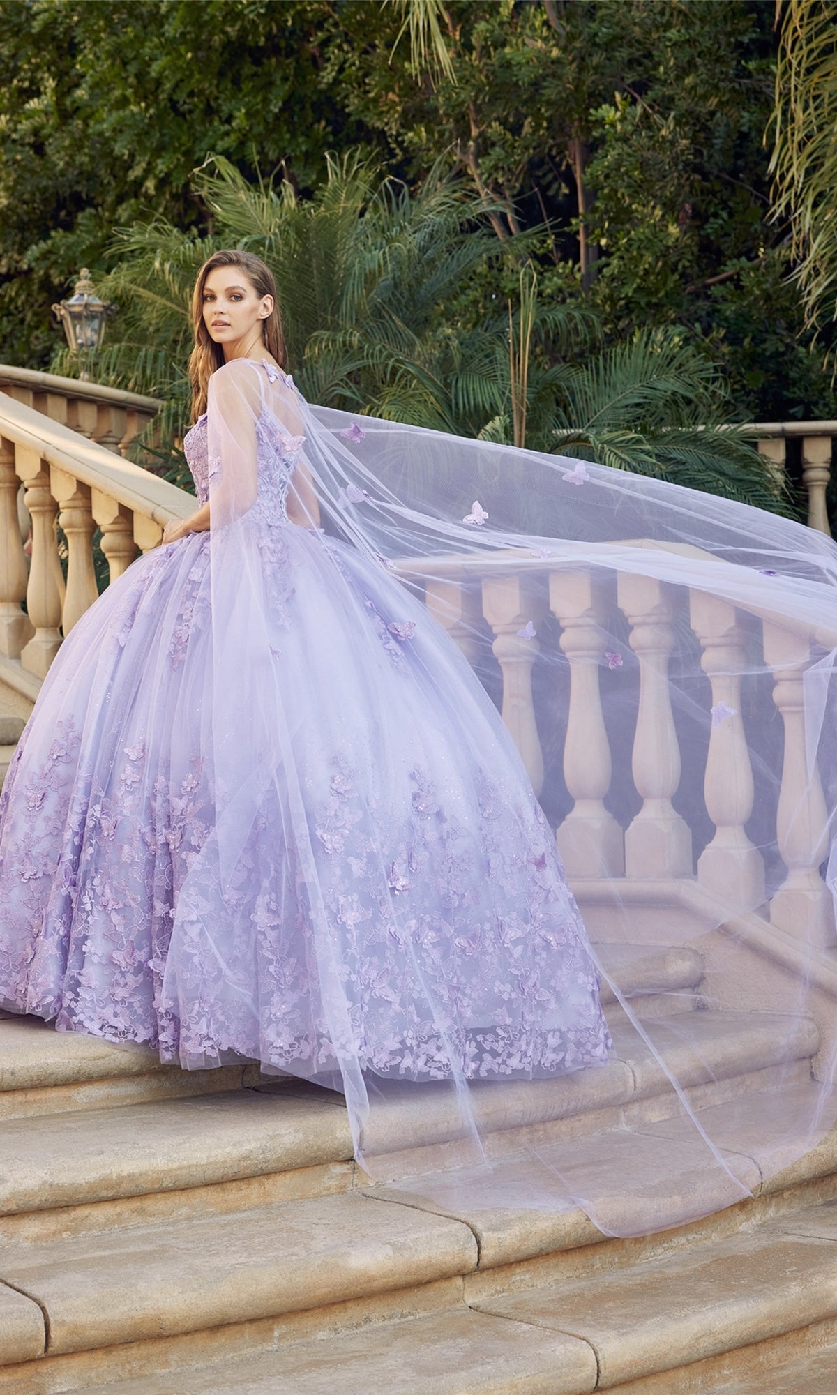 A woman in the Juliet 1442 Quinceañera Dress with Sheer Cape, featuring lavender embroidery and floral details, stands on stone steps before a balustrade amid greenery.